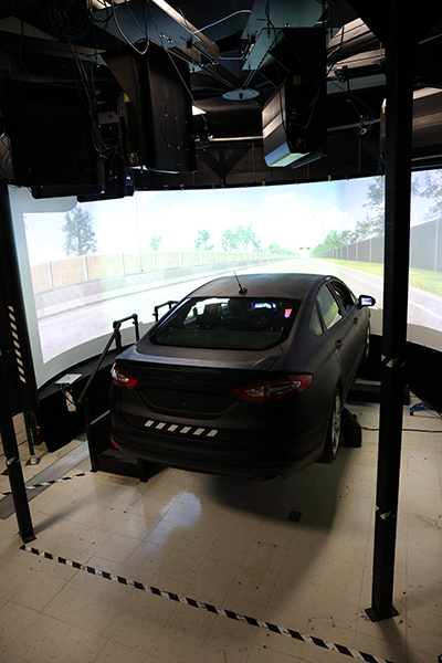 An elevated sedan is partially encircled by a simulated highway scene projected on a set of screens. 