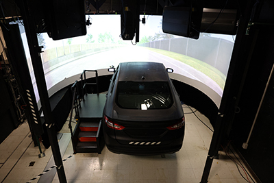 An elevated sedan chassis is placed in front of a screen showing a simulated highway scene.