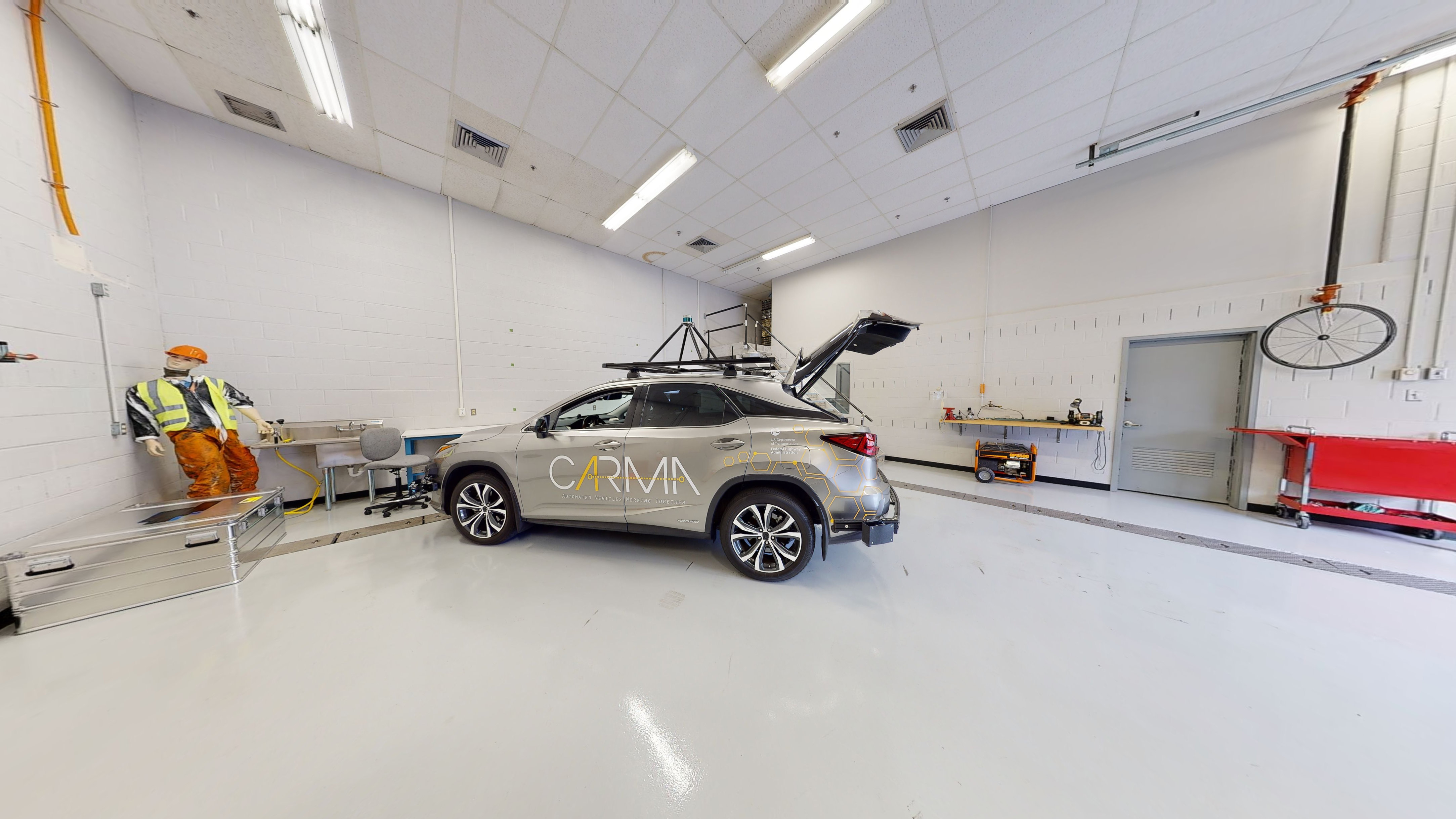 Part of a garage space with a car with equipment on its roof that supports self-driving parked in front of a test dummy wearing a hard hat, safety vest, and work pants.