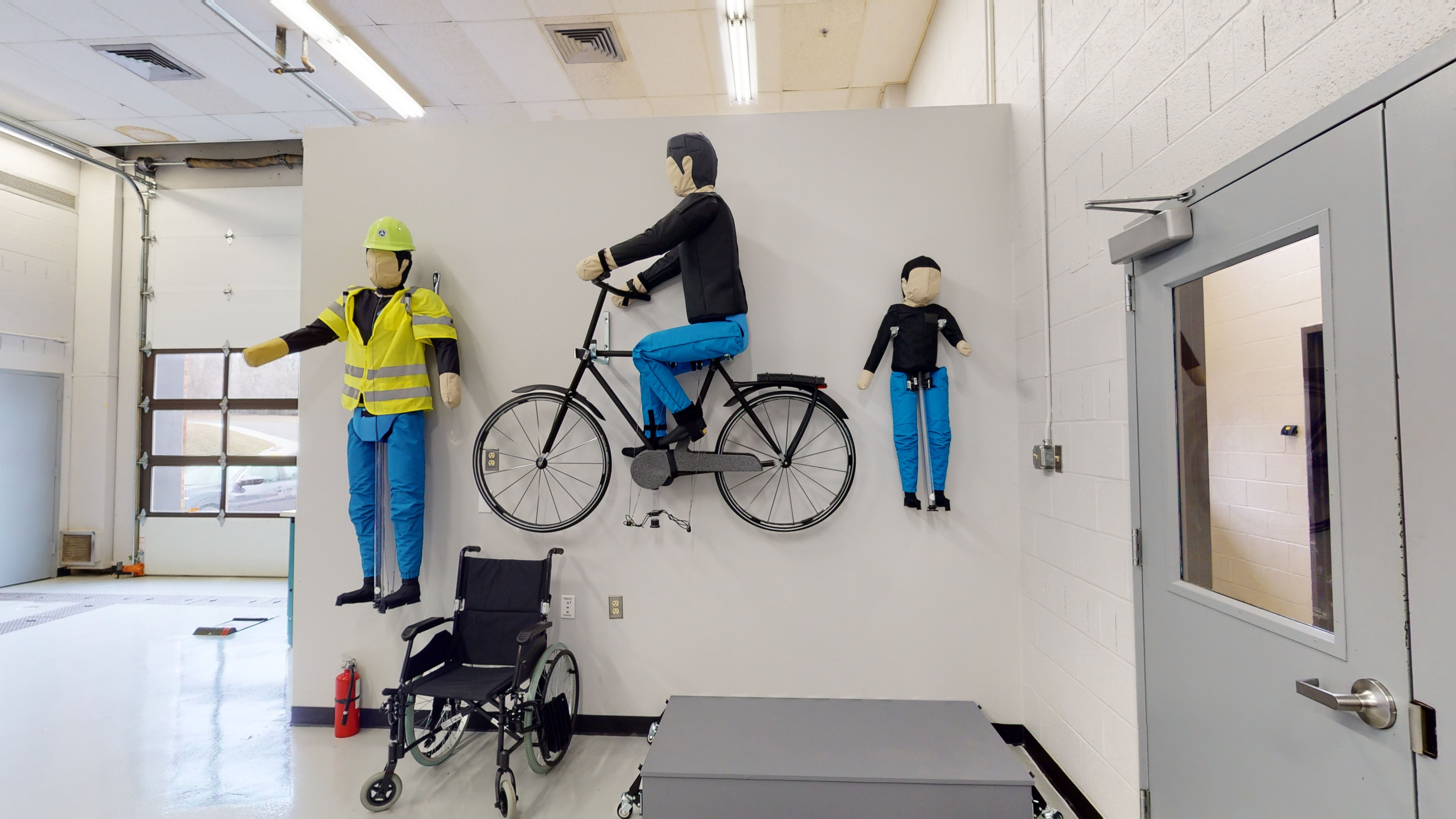 Three test dummies are mounted on a wall in a garage space. The far-left dummy is wearing a hard hat, safety vest, and pants. The test dummy in the middle is on a bicycle. The far-right dummy appears to be the size of a child.