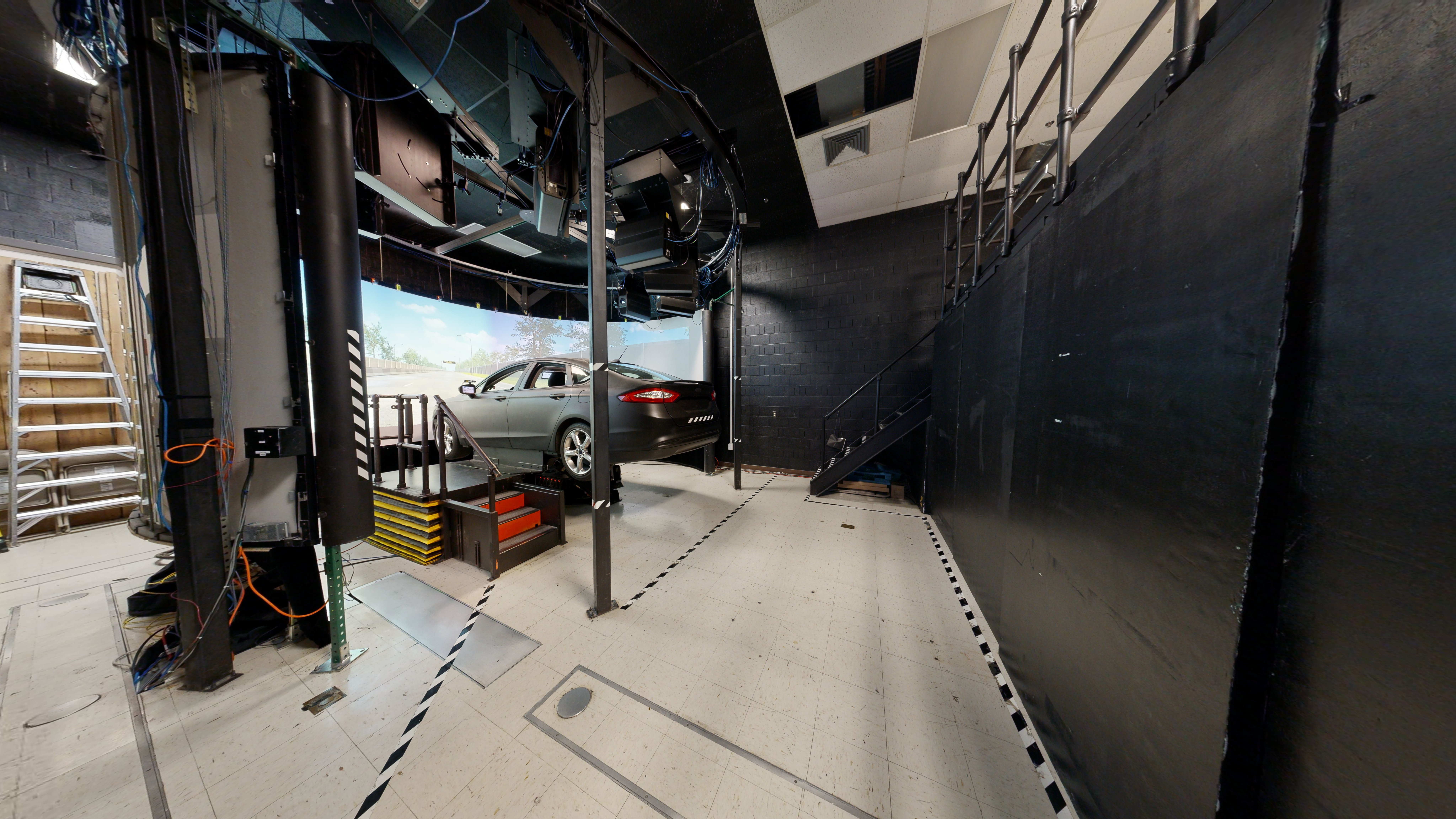 A room with a sedan in front of a screen displaying a simulated highway scene. 