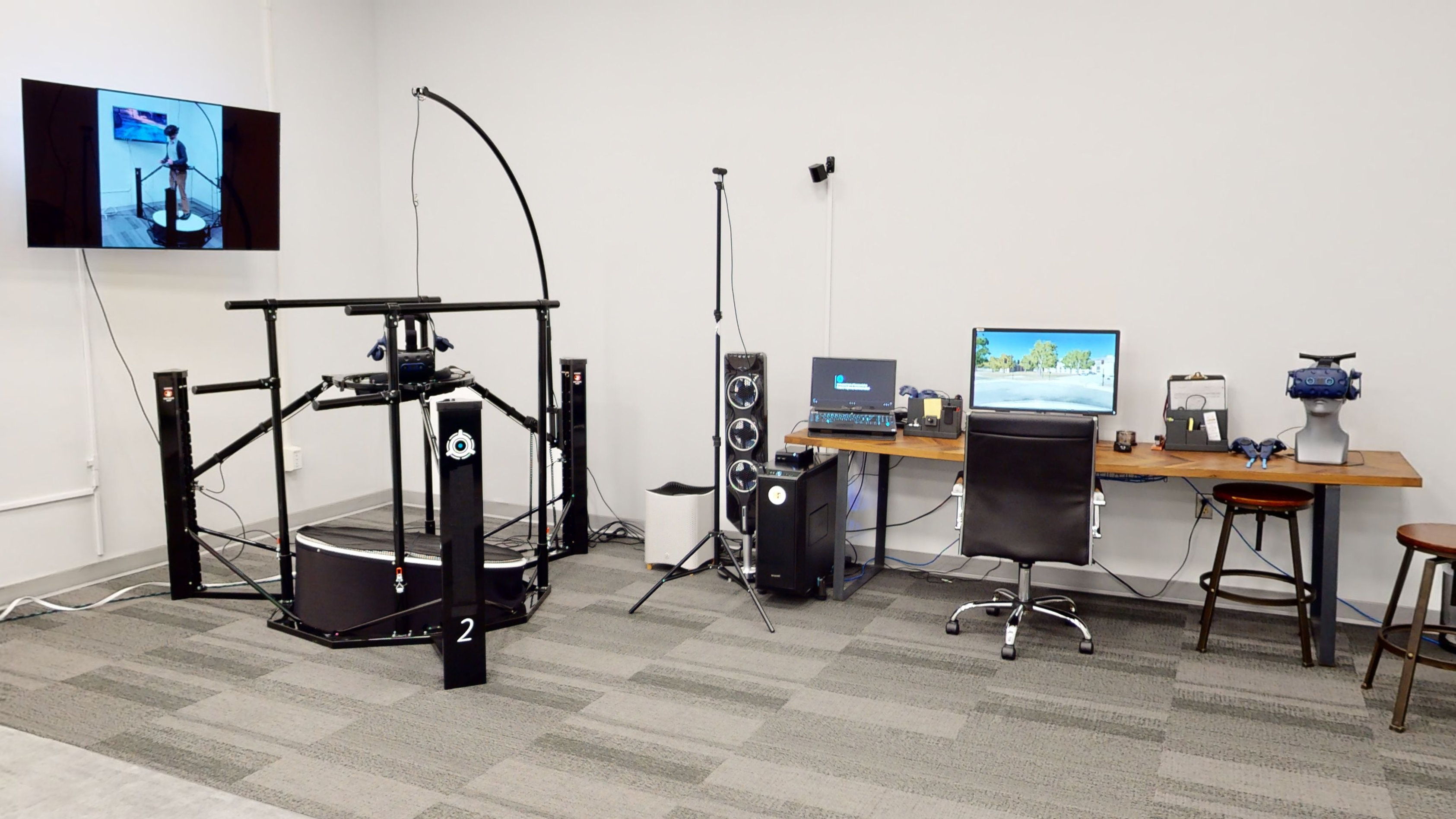 An omnidirectional treadmill, headset, mounted television screen, and motion-tracking cameras next to a computer workstation, a virtual reality headset, and associated equipment.