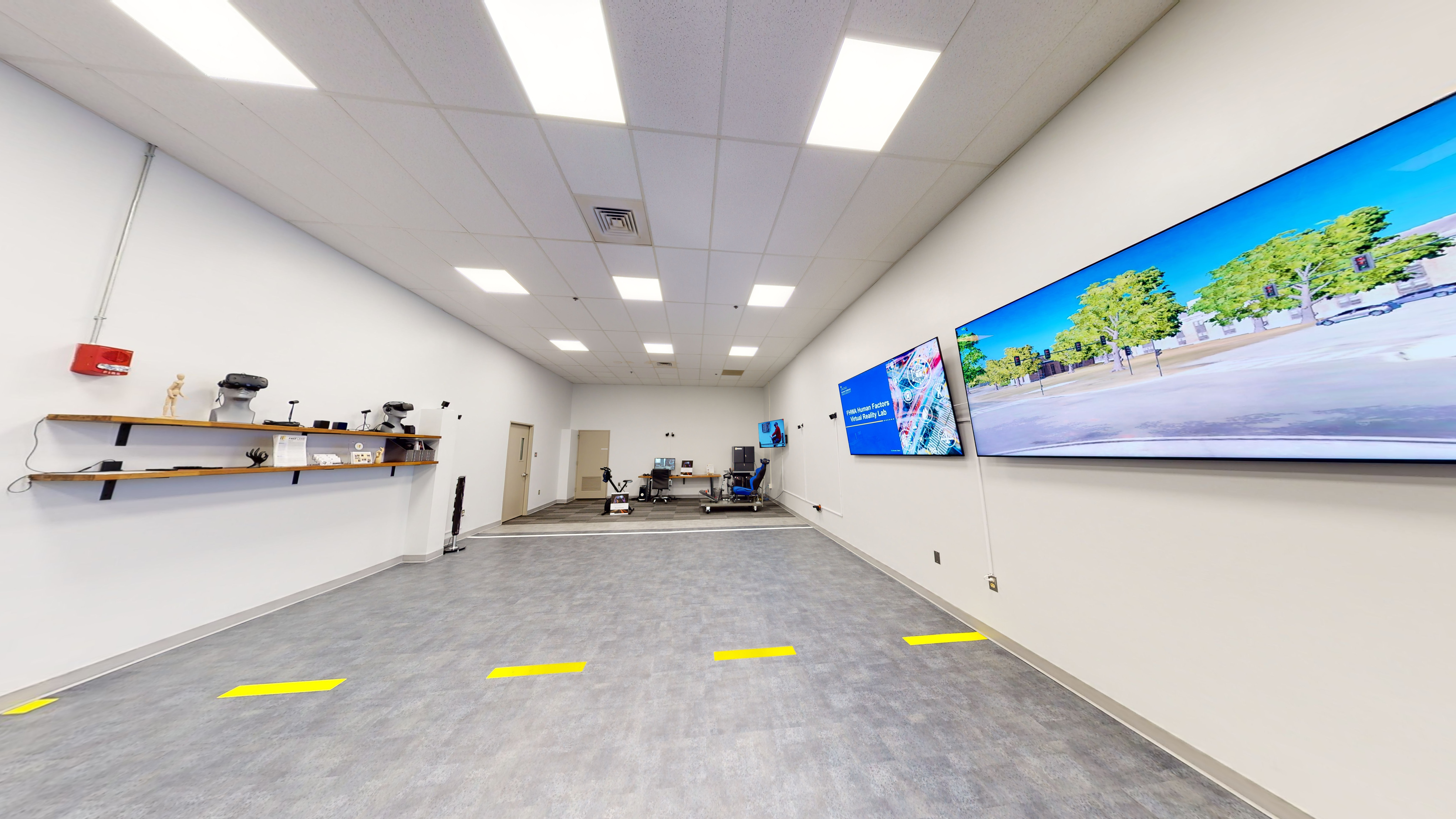 A room for virtual reality testing that includes several wall-mounted screens, virtual reality headsets, mounted motion-tracking cameras, a computer workstation, a virtual reality bicycle simulator, and a vehicle-driving simulator. 