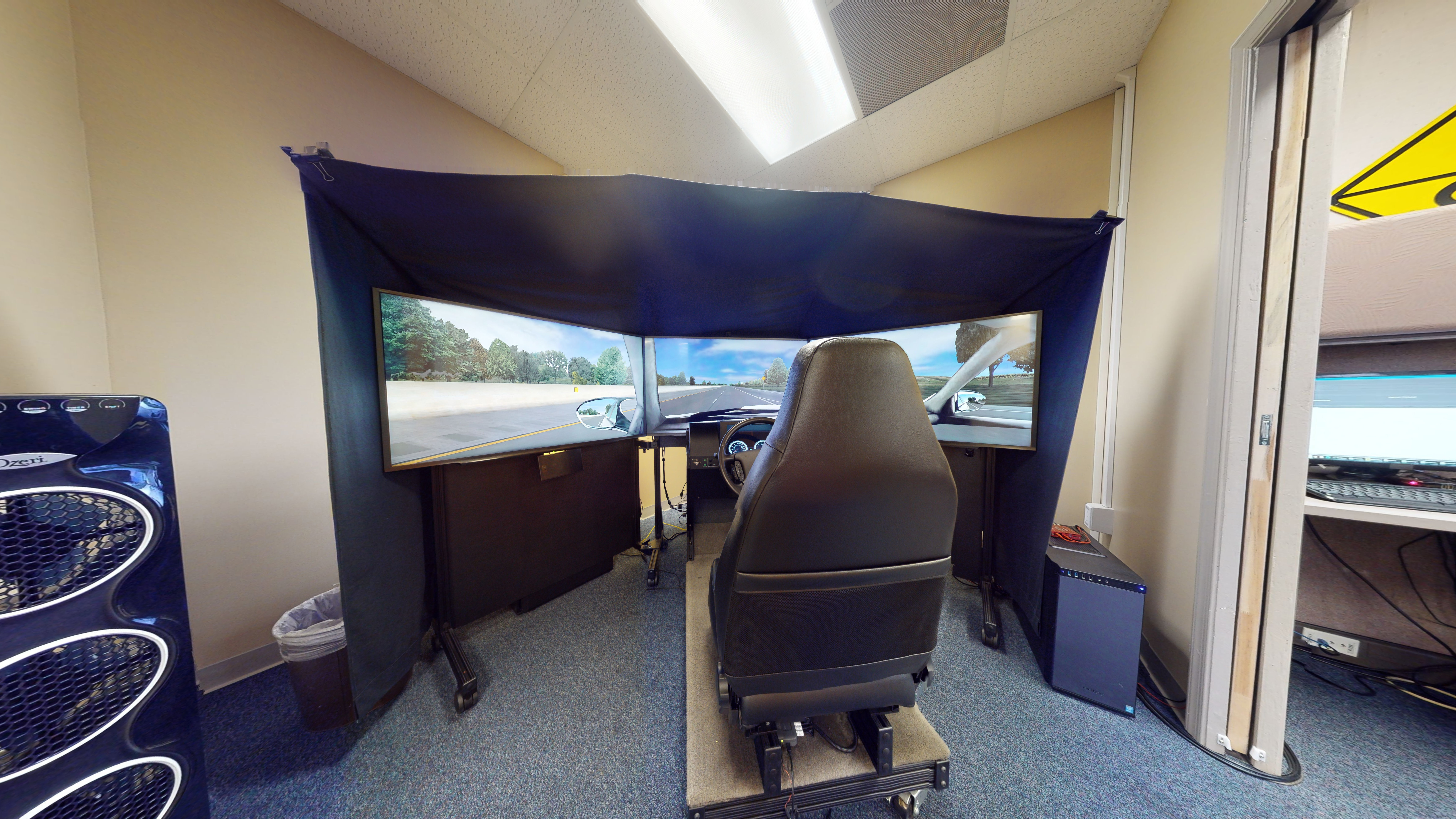 A driving simulator set up that includes three monitors positioned in a half circle in front of a seat and a steering wheel. 