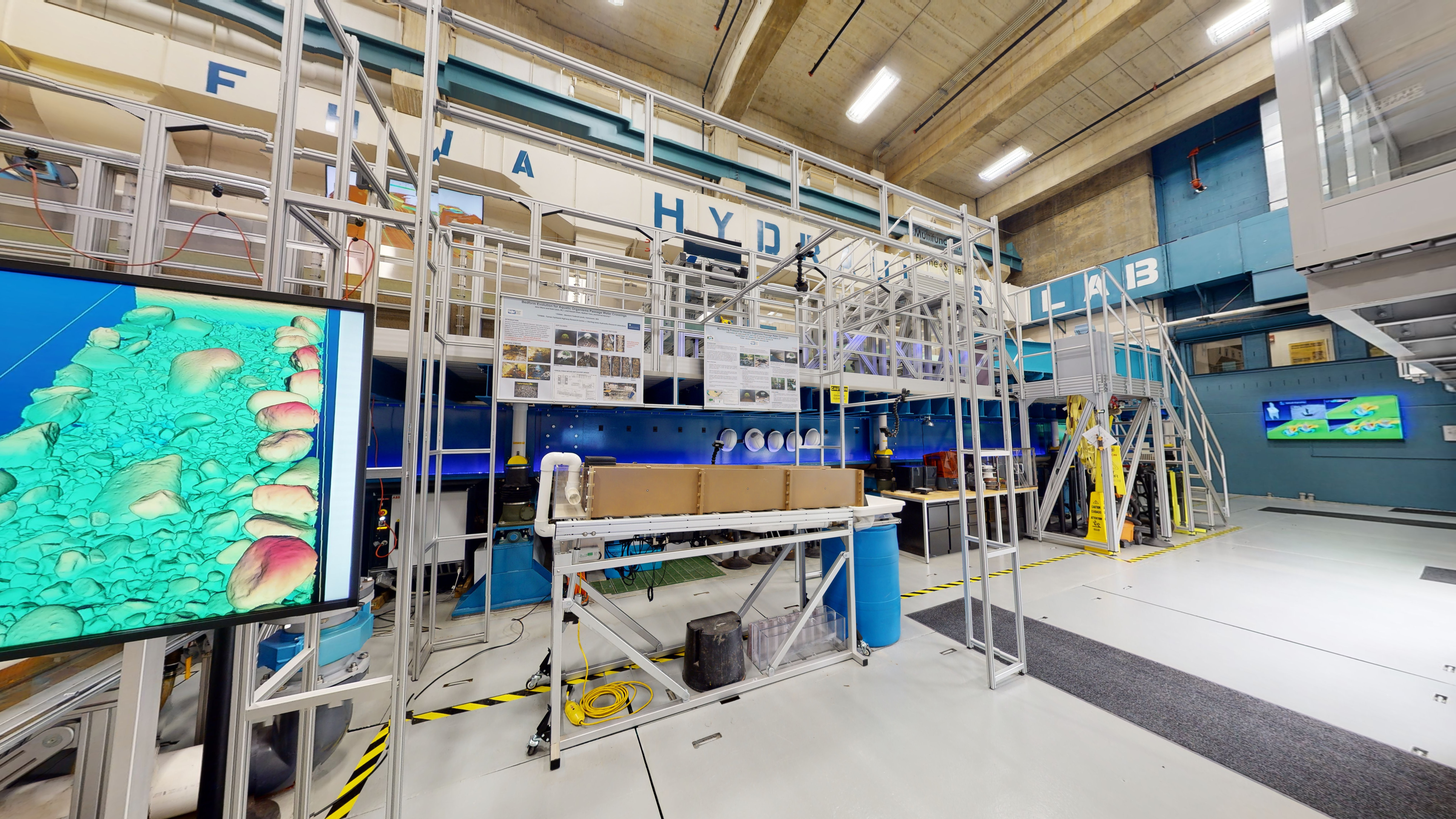 Ground floor of a laboratory facing the flume system containing tables, cabinets, hard hats, caution signs, straps, and other various tools, equipment, devices, and a monitor.  