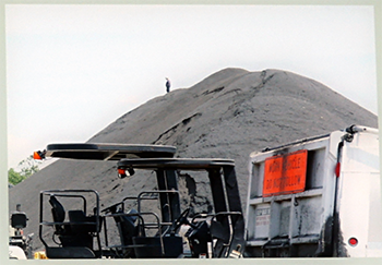 A large stockpile of reclaimed asphalt pavement rises above several vehicles and a dump truck.  