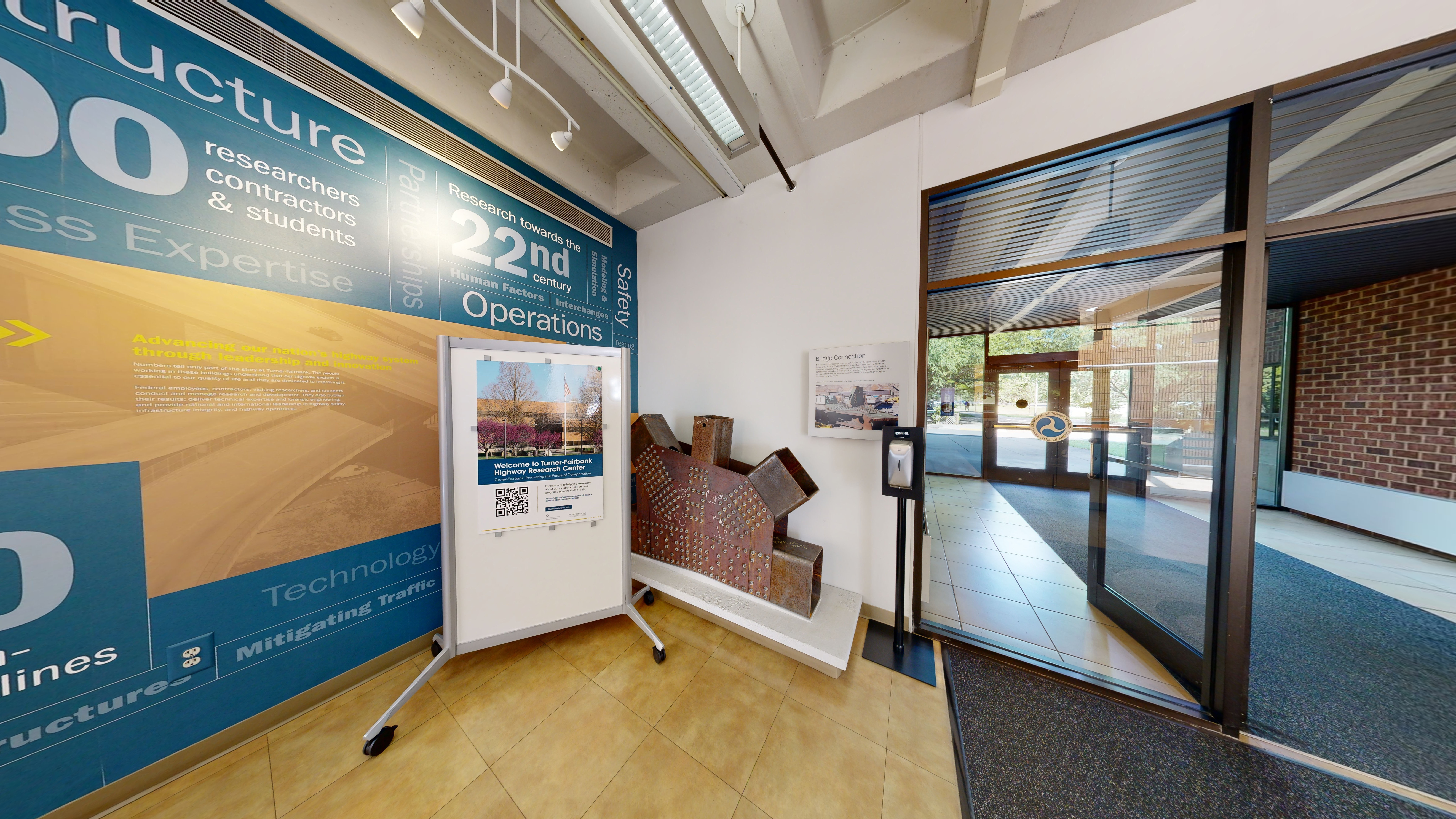 A large text and photograph-based wall decoration on the left that features facts and figures about the Turner-Fairbank Highway Research Center. A sign in front of the wall includes a photograph of the building exterior, welcomes visitors to the center, and features a QR code for more information about the site. 