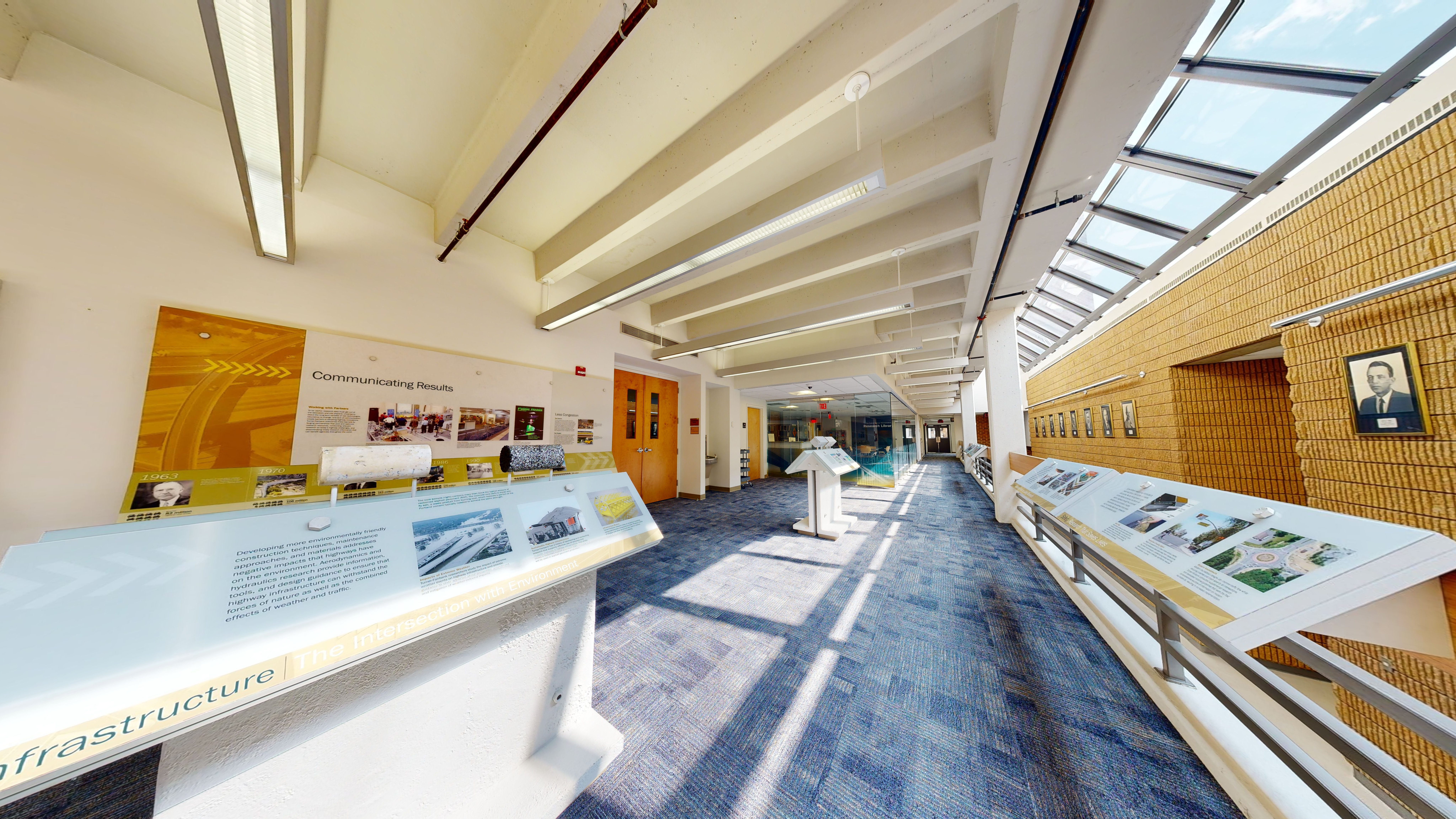 Hallway filled with informational signage and portraits on the walls and right-side railing. Two standalone informational displays are in the center of the hallway. 