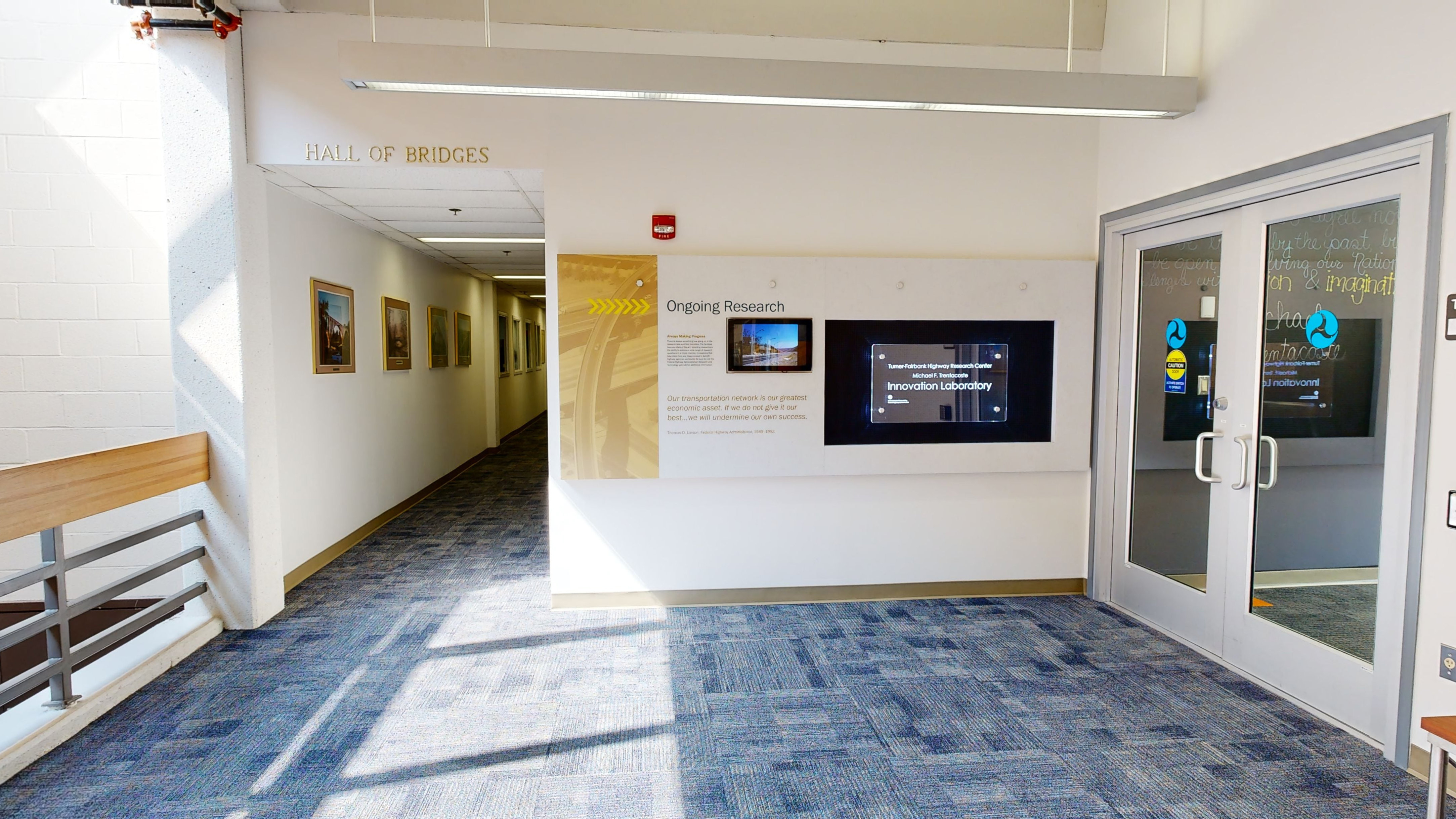 Area leading to a hallway on the left labeled “Hall of Bridges” with an informational display on a wall perpendicular to a set of double doors. 