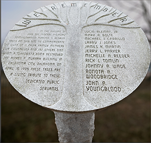 A circular memorial plaque contains the image of a tree with the words “we remember.” The plaque states: “In the autumn of 1995, the Federal Highway Administration planted 11 redbud trees on this site to commemorate the lives of 11 FHWA family members. Our colleagues and 157 others died when a terrorist bomb destroyed the Alfred P. Murrah building in Oklahoma City, Oklahoma, on April 19, 1995. These trees are a living tribute to these dedicated public servants. Lucio Aleman, Jr., Mark A. Bolte, Michael J. Carrillo, Larry J. Jones, James K. Martin, Jerry L. Parker, Michelle A. Reeder, Rick L. Tomlin, Johnny A. Wade, Ronata A. Woodbridge, John A. Youngblood.”