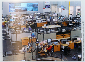 A large room filled with cubicles, desks, and computers with multiple wall-mounted monitors displaying highway camera feeds.