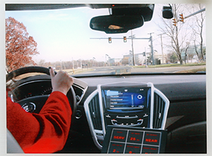 The interior of a car with a driver behind a steering wheel approaching a traffic signal intersection.