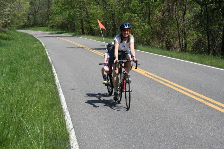 Person and child wearing helmets riding a tandem bicycle with a safety flag on a two-lane highway surrounded by grass and forest
