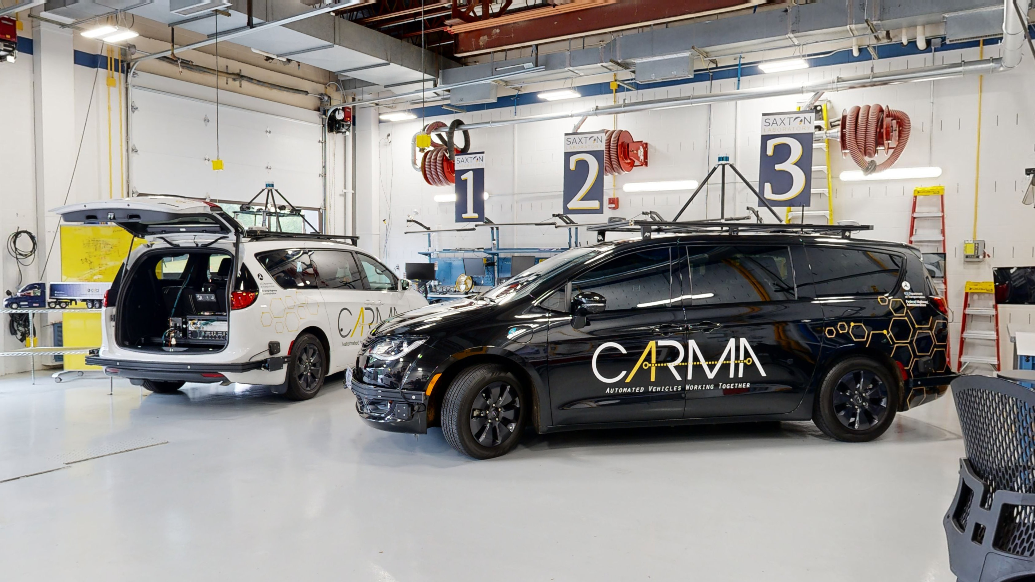 Two vans with equipment installed on their respective roofs that support self-driving. The open trunk of the left van reveals additional equipment to support self-driving functions.