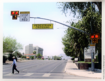 A pedestrian crosswalk with a traffic light on a pole hanging over it that has the signs “Crosswalk: Stop on Red” and “Pedestrian Crossing.” At the street corner next to the crosswalk, is a vertical pole with traffic signals and a sign “Crosswalk: Stop on Red.” 