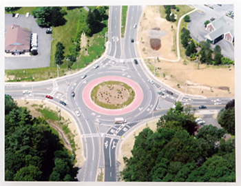 An aerial view of a traffic roundabout nestled between two tree-filled areas and two sets of buildings.