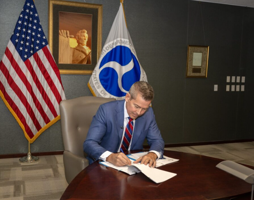 Sean Duffy seated at a desk signing papers with the U.S. and U.S. Department of Transportation flags and an image of the Lincoln Memorial on the wall behind him.