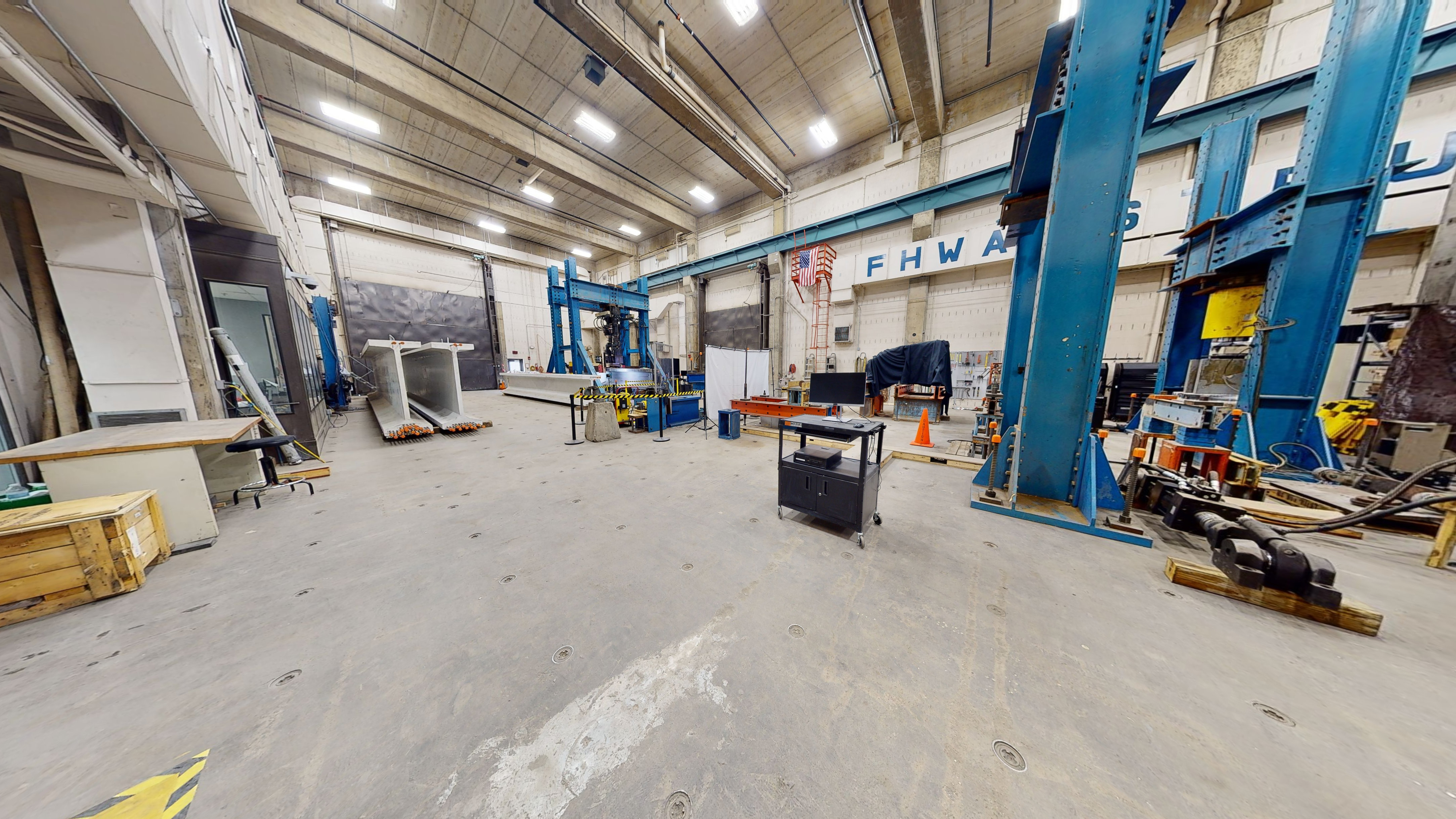 Laboratory space filled with an assortment of equipment for testing the performance of bridge structures and structural systems, including large-scale beam static testing frames, servo-hydraulic actuators, a high-force vertical load frame outfitted with a high-force hydraulic ram, the large-scale beam fatigue test and the strand development length test setup, and two tall concrete girders with embedded prestressing strands. 