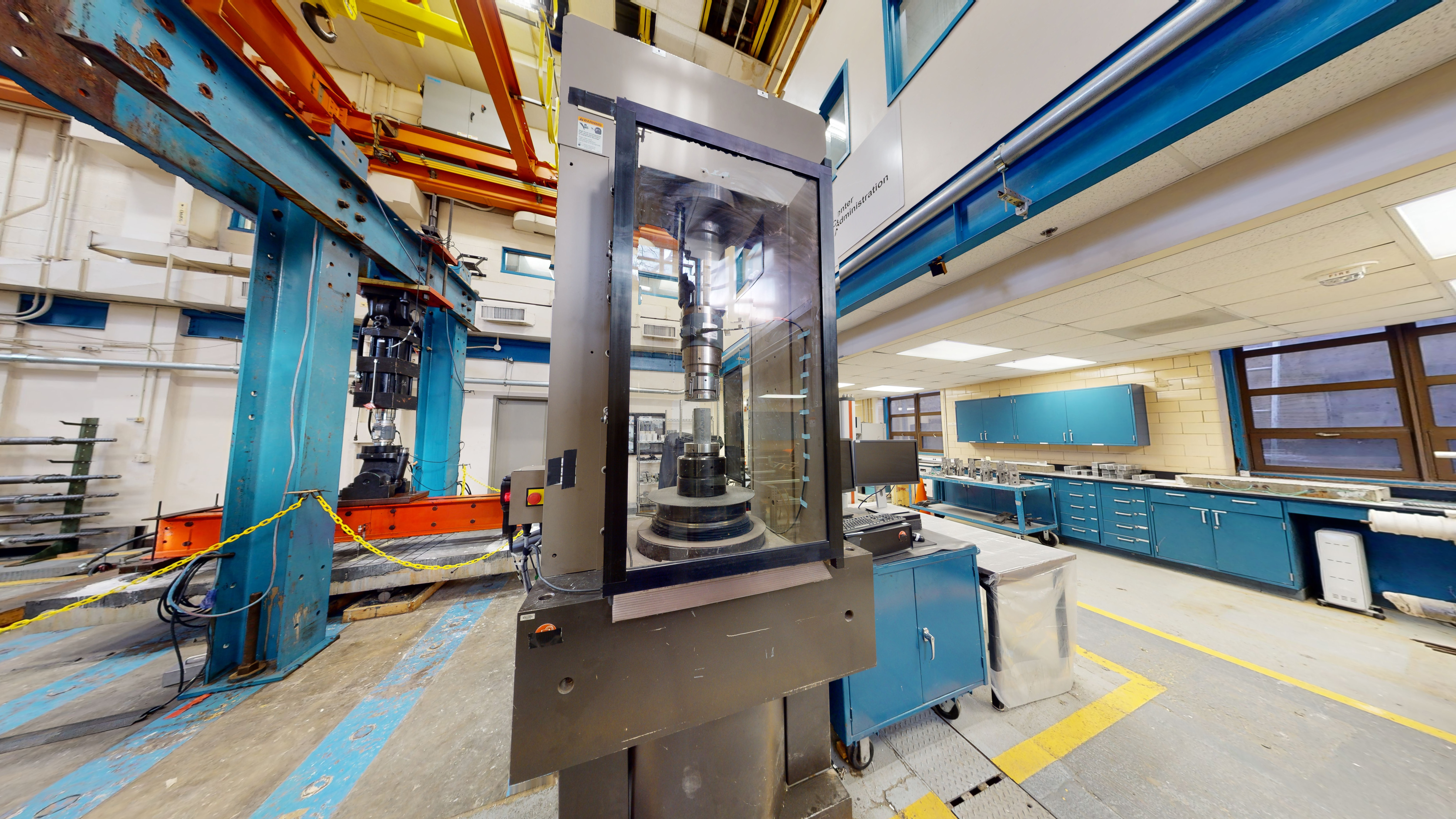 Part of a laboratory space with a high-capacity compression testing machine, a structural reaction frame with an actuator and specimen, and cabinets on the right side.