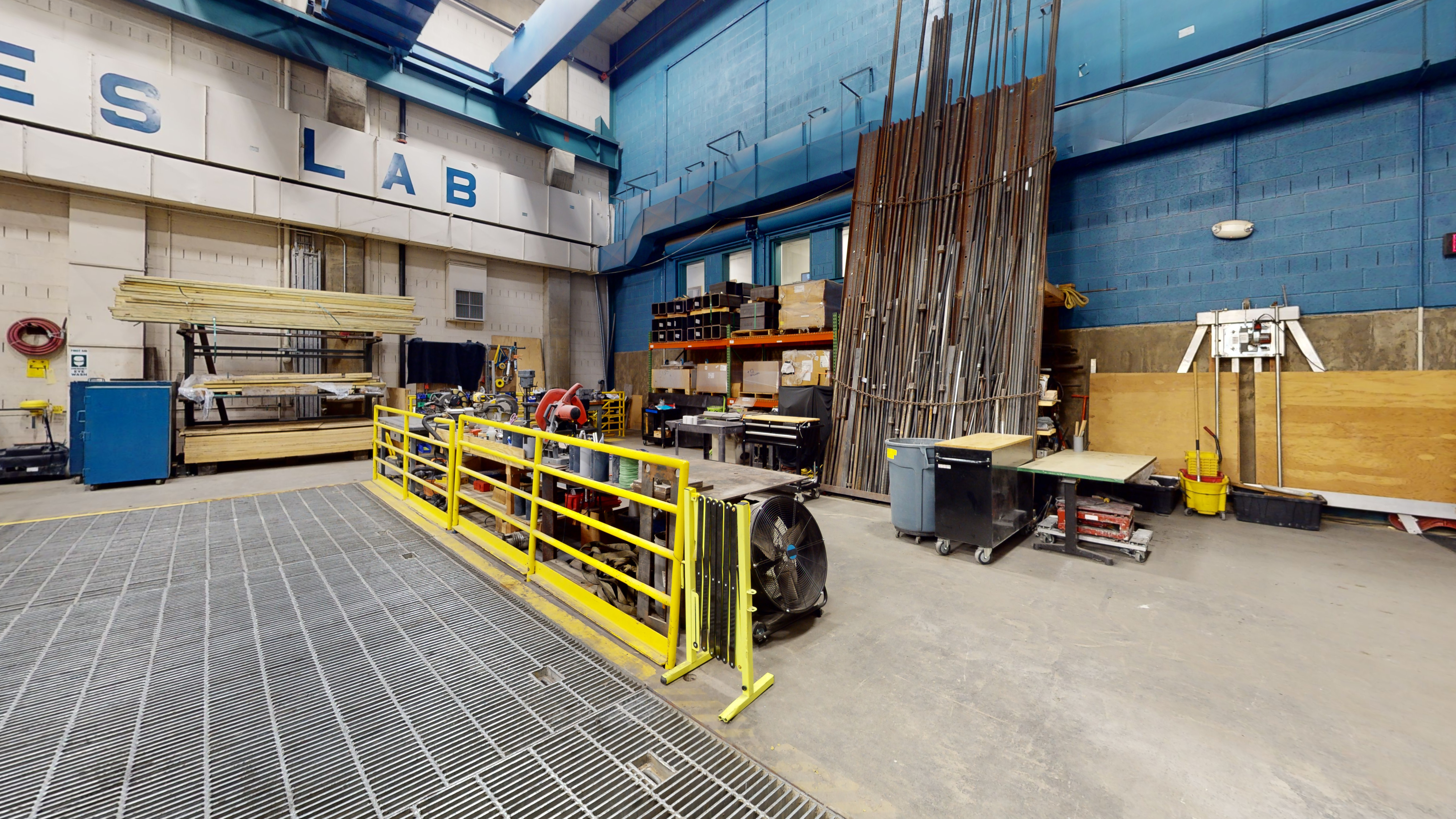 Part of a laboratory space filled with metal floor grates, tall pieces of slender metal material, and different sizes and lengths of wood and plywood stacked on shelves on the wall.  