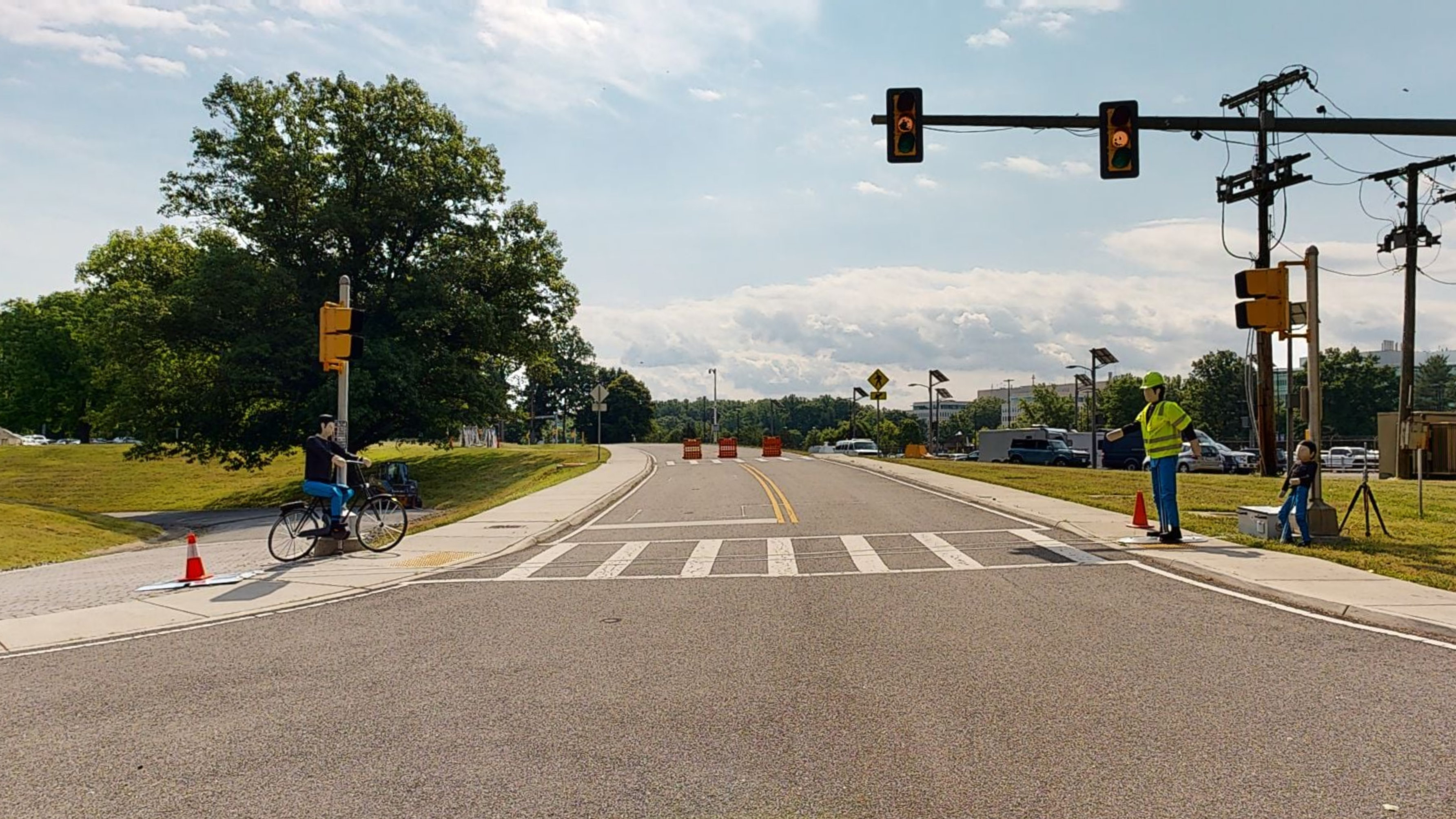 A street intersection with traffic and pedestrian walk signals, a crosswalk, and two test dummies (one on a bicycle and one wearing a hard hat and safety vest).