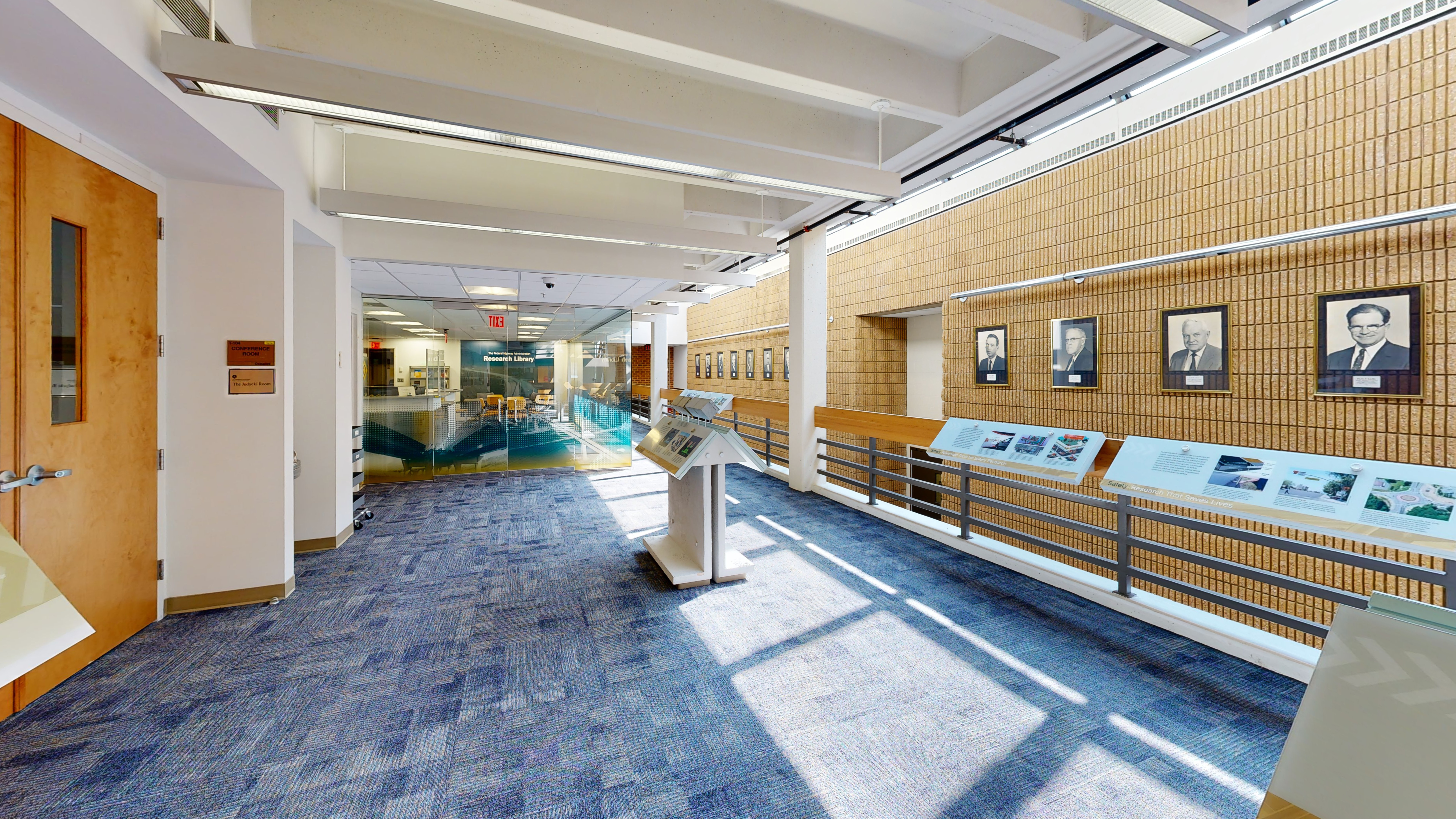 A wide overlook area is decorated with informational signage and portraits hung on a brick wall. In the center of the wide hallway is an information stand. 