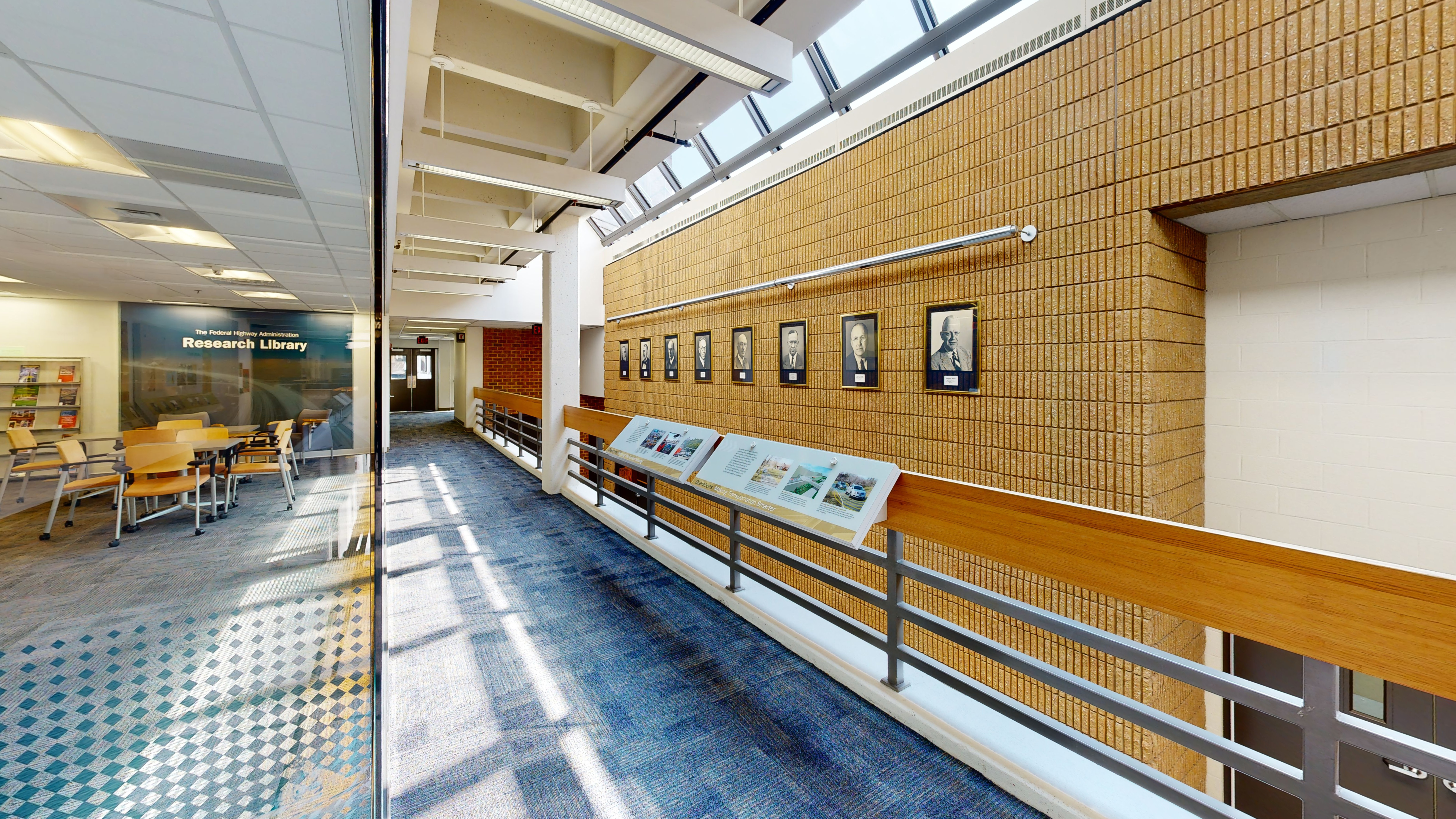 Hallway with a room to the left with chairs and a bookshelf and, on the right, portraits on a brick wall, and informational signage on a railing.