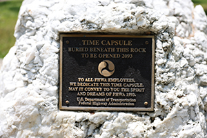 A stone  with a plaque shows the logo of the U.S. Department of Transportation and states, “Time capsule buried beneath this rock to be opened 2093. To all FHWA employees, we dedicate this time capsule. May it convey to you the spirit and dreams of FHWA 1993. U.S. Department of Transportation Federal Highway Administration.”