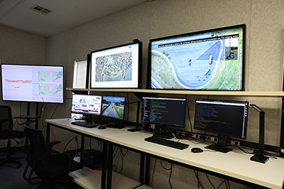 Several monitors and computers on desks display aerial photographs, code, roadways, and graphs.