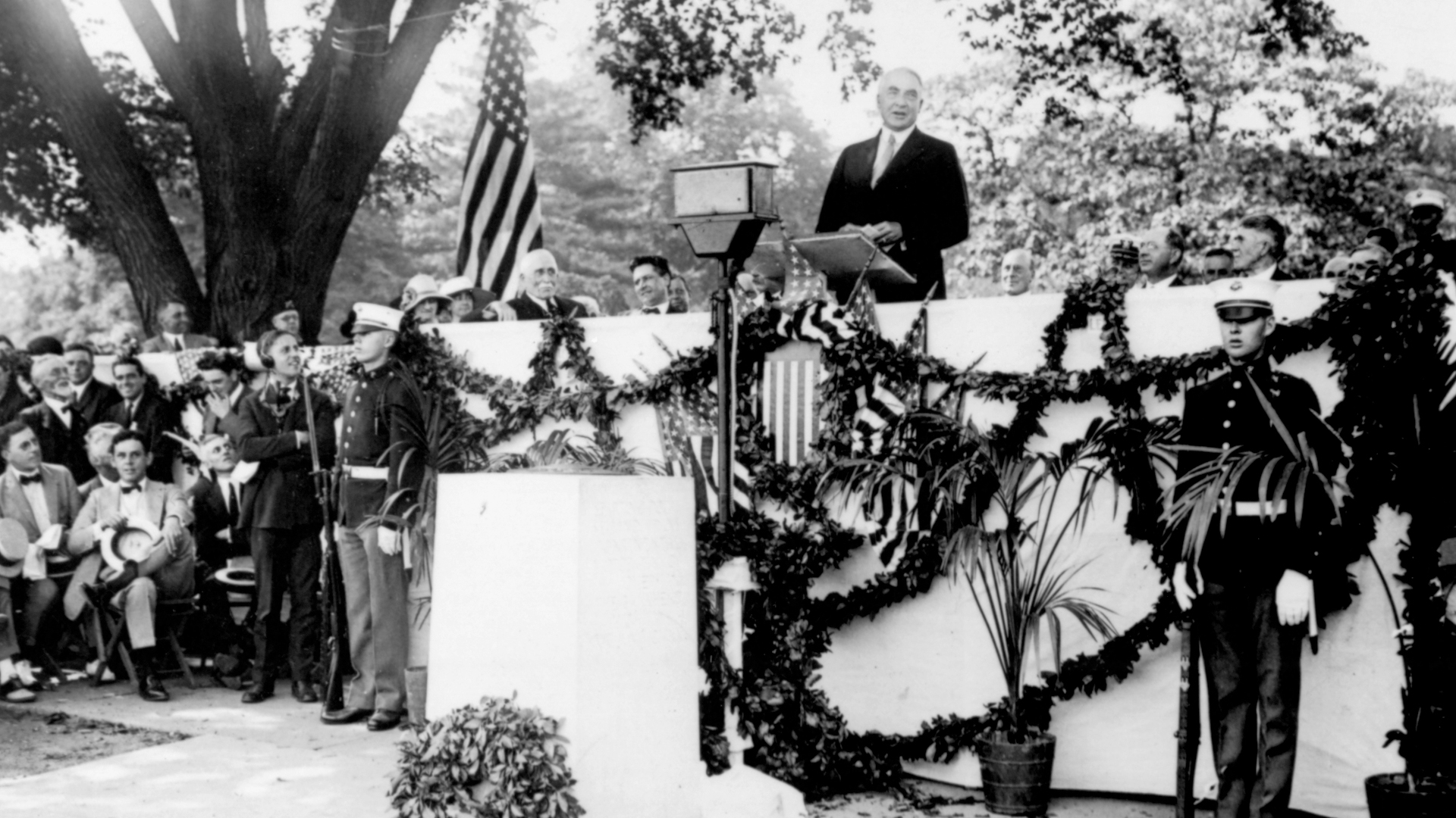 President Harding stands at a podium delivering a speech. The stage is adorned with garland and U.S. flags. A crowd is gathered, and military personnel stand nearby.